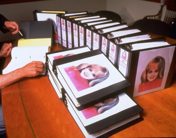 Murdered 6-yr-old beauty queen JonBenet Ramsey's image amblazoned on dozens of binders crammed w. investigators's reports for case which still unsolved after 6 months, in Sheriff's office. 