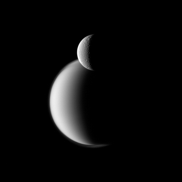 Craters appear well defined on icy Rhea in front of the hazy orb of the much larger moon Titan in this Cassini spacecraft view of these two Saturn moons.