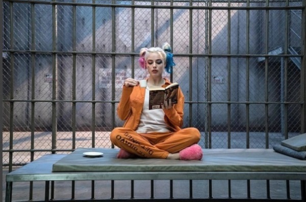 Harley Quinn is comfortably sitting, reading, and drinking coffee inside her cell.